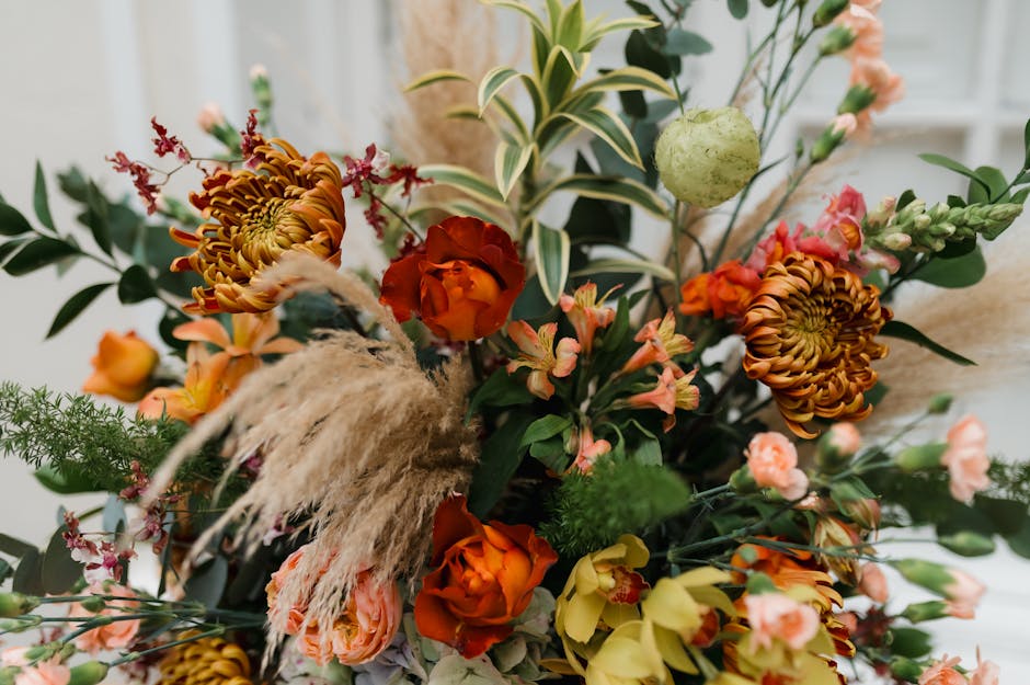 A vibrant floral arrangement featuring a mix of orange, yellow, and pink blooms including chrysanthemums, roses, and small snapdragons, complemented by lush green foliage with variegated leaves. The b