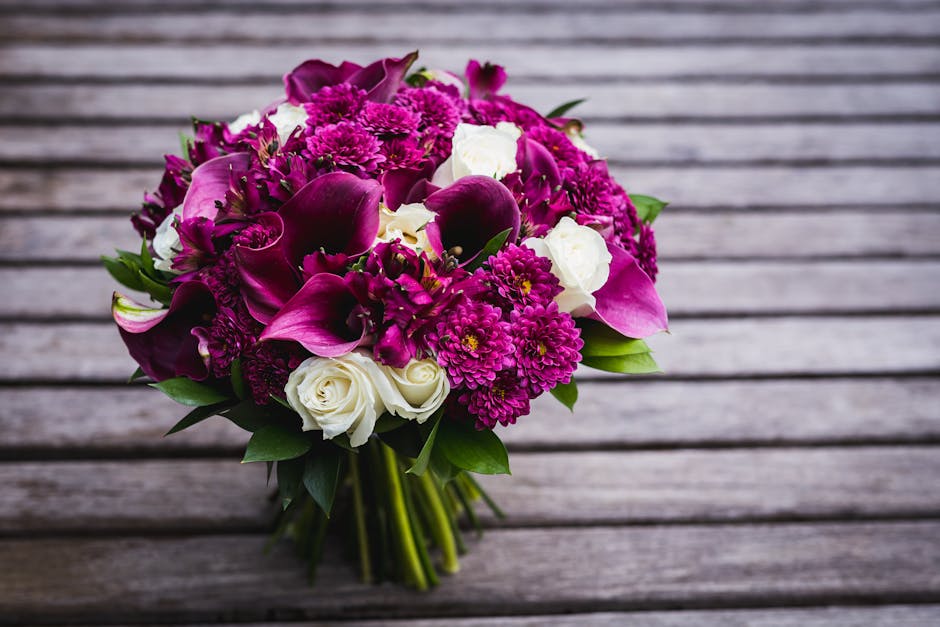 A vibrant bouquet of fresh flowers arranged with deep purple calla lilies, bright magenta chrysanthemums, and delicate white roses, all intertwined with lush green foliage. The bouquet is tied with a