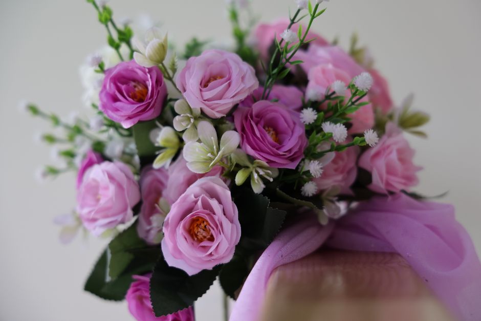 A floral arrangement featuring a bouquet of fresh pink roses with delicate, layered petals, complemented by small white daisies and light green foliage. The flowers are wrapped in soft pink tissue pap